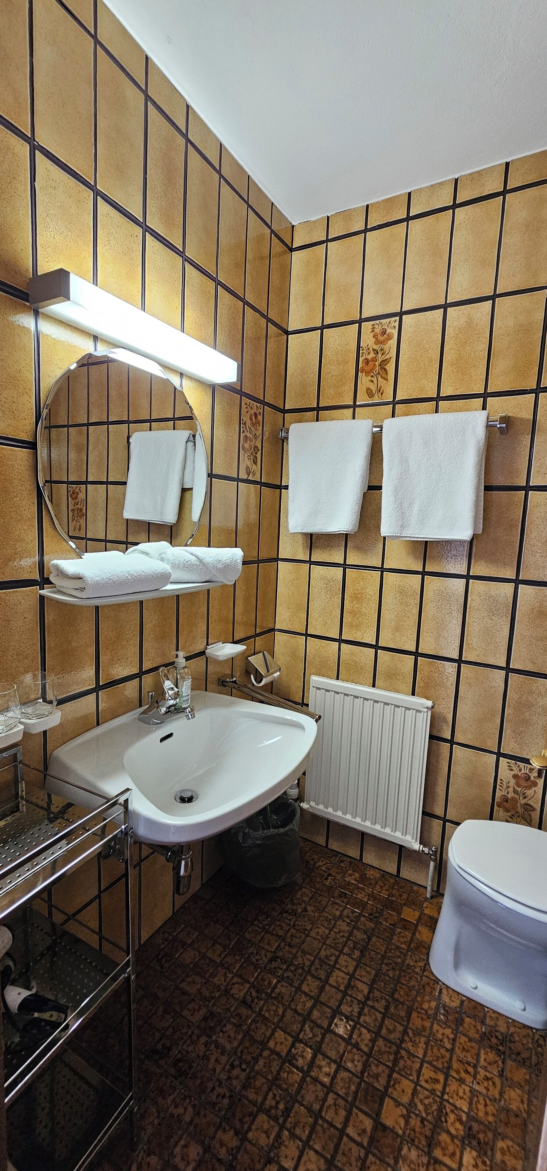 Ein Badezimmer mit einem weißen Waschbecken, einem Spiegel und einer Toilette. Zwei Handtücher hängen an der Wand.