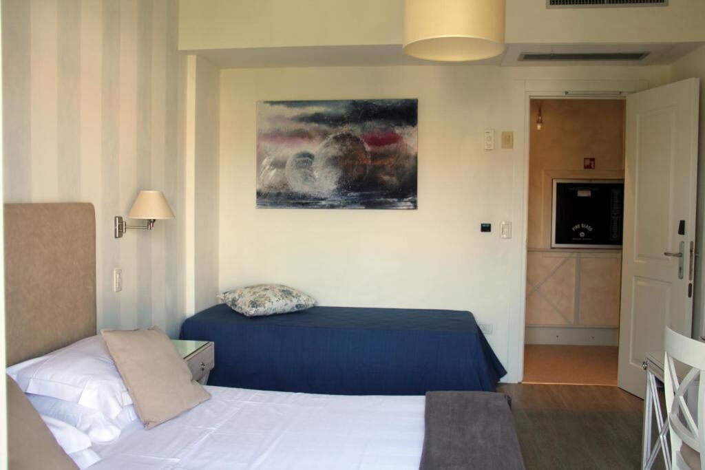 A hotel room with a bed, a nightstand, a lamp, a painting on the wall, and a kitchenette in the background.