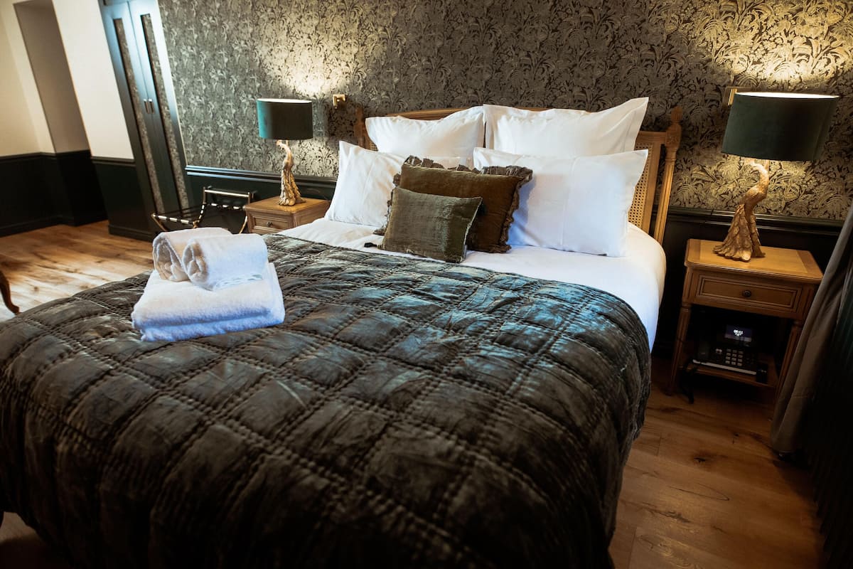 Un lit bien fait, avec une couvre-lit matelassé, plusieurs oreillers et une serviette pliée posée sur le côté. On y trouve deux tables de chevet avec des lampes, une tête de lit en bois et un papier peint à motifs.