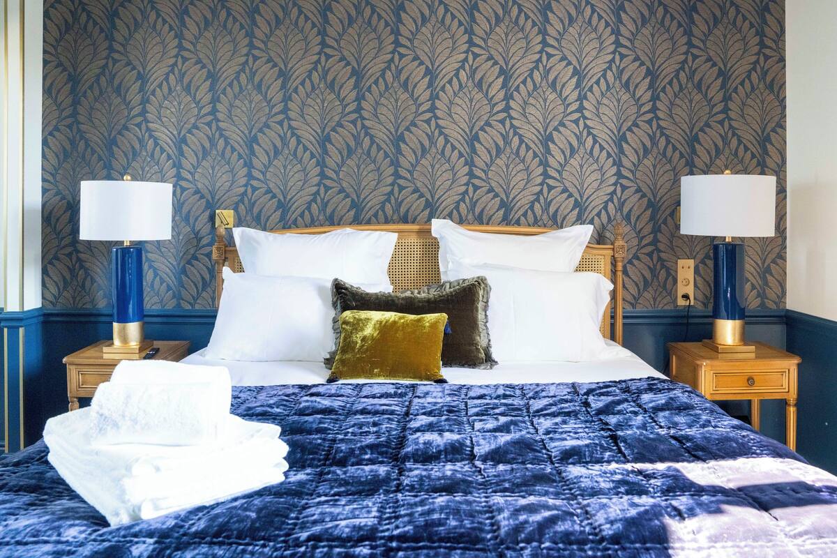 Un lit bien fait, recouvert d’une courtepointe bleue, avec des oreillers blancs et dorés, et une tête de lit dorée. On y trouve deux tables de chevet avec des lampes, et un mur orné d’un motif végétal.