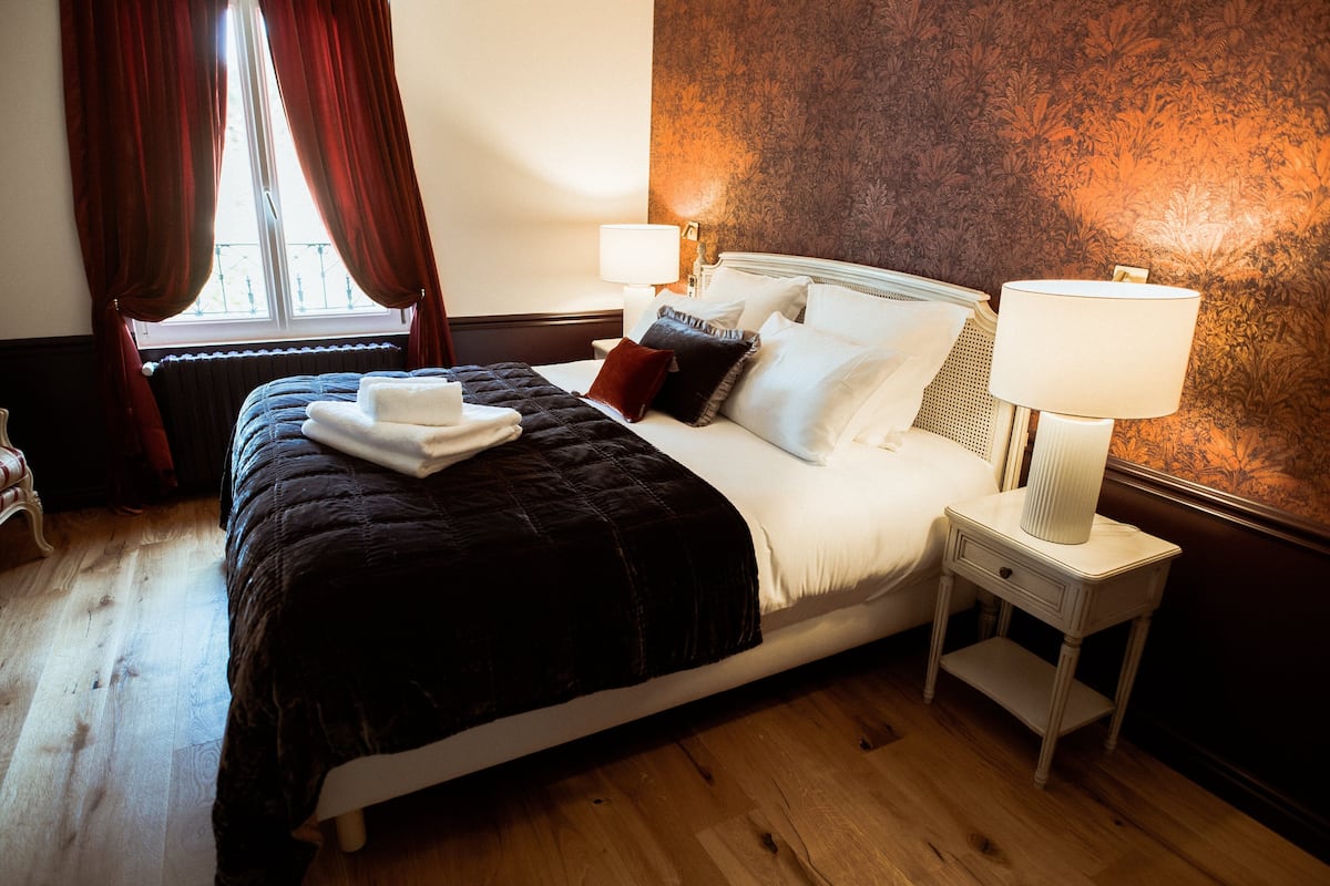 Un lit bien fait, recouvert d’une couverture sombre, avec du linge de lit blanc et un oreiller rouge. Une table de chevet, sur laquelle se trouve une lampe et un petit tiroir, est placée à côté du lit. La pièce est dotée d’un plancher en bois et d’une fenêtre ornée de rideaux rouges.