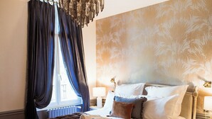 Ein Schlafzimmer mit einem großen Bett, einer Kristallleuchtre, einem Fenster mit Vorhängen und einem Nachttisch mit Lampe.