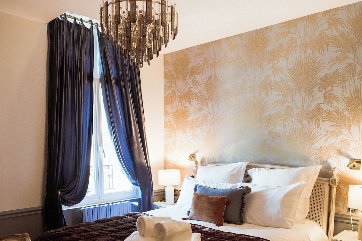 Une chambre à coucher avec un grand lit, un lustre, une fenêtre avec des rideaux et une table de chevet avec une lampe.