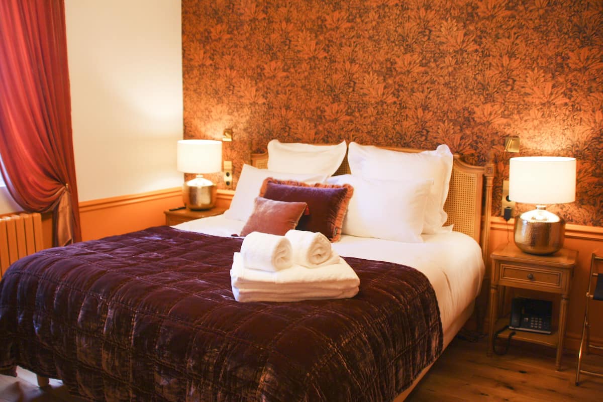 Un lit bien fait, recouvert d’une couverture sombre, avec deux lampes de chevet et une pile de serviettes pliées posée sur le lit.