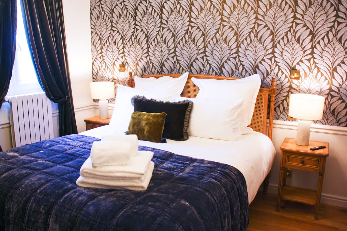 Un lit bien fait, avec une couverture bleue, des oreillers blancs et une serviette blanche pliée. Il y a deux tables de chevet avec des lampes et un mur orné d’un motif de feuilles.