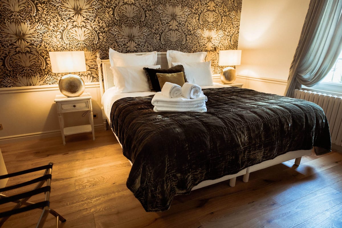 Un lit bien fait, recouvert d’une couverture sombre, avec des oreillers blancs et une serviette pliée posée sur le côté. On y trouve deux tables de chevet avec des lampes et un motif décoratif sur le mur.