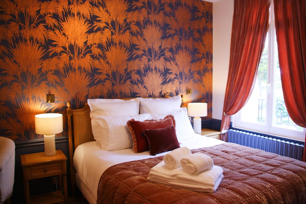 Une chambre d’hôtel avec un grand lit, deux tables de chevet avec des lampes, une fenêtre avec des rideaux rouges et un papier peint à motifs floraux.