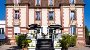 Ein großes, mehrgeschossiges Gebäude mit einer symmetrischen Fassade, das einen zentralen Eingang mit Stufen, zwei schwarze Sonnenschirme und Pflanzkübel ziert.