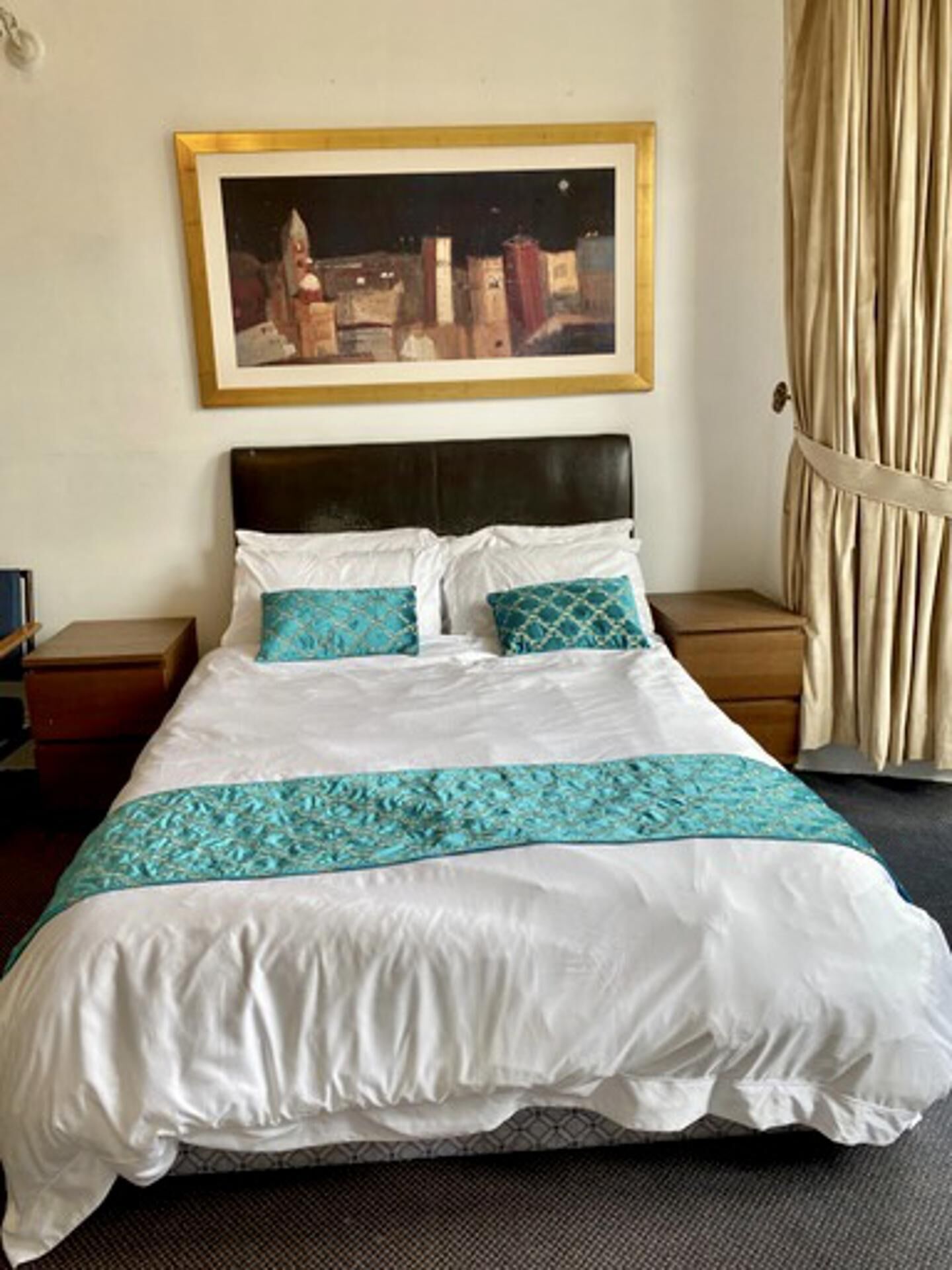 A neatly made bed with a white and turquoise bedspread, a dark headboard, and a framed cityscape painting above it.