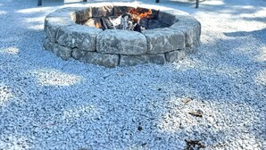 A fire pit with two Adirondack chairs, surrounded by gravel, with a house and trees in the background.