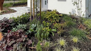Una casa moderna con un jardín cuidado, una pérgola de madera y una casa roja al fondo.