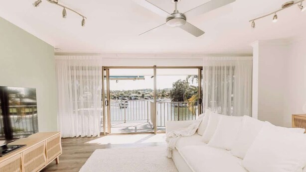 A modern living room with a white sofa, a wooden TV stand, and a large window with a view of a cityscape. - 'Key Point' - Tranquil Getaway - Mooloolaba (Mooloolaba)