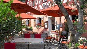 Un café al aire libre con mesas de madera y sombrillas rojas, rodeado de plantas en macetas y una fachada de edificio tradicional.