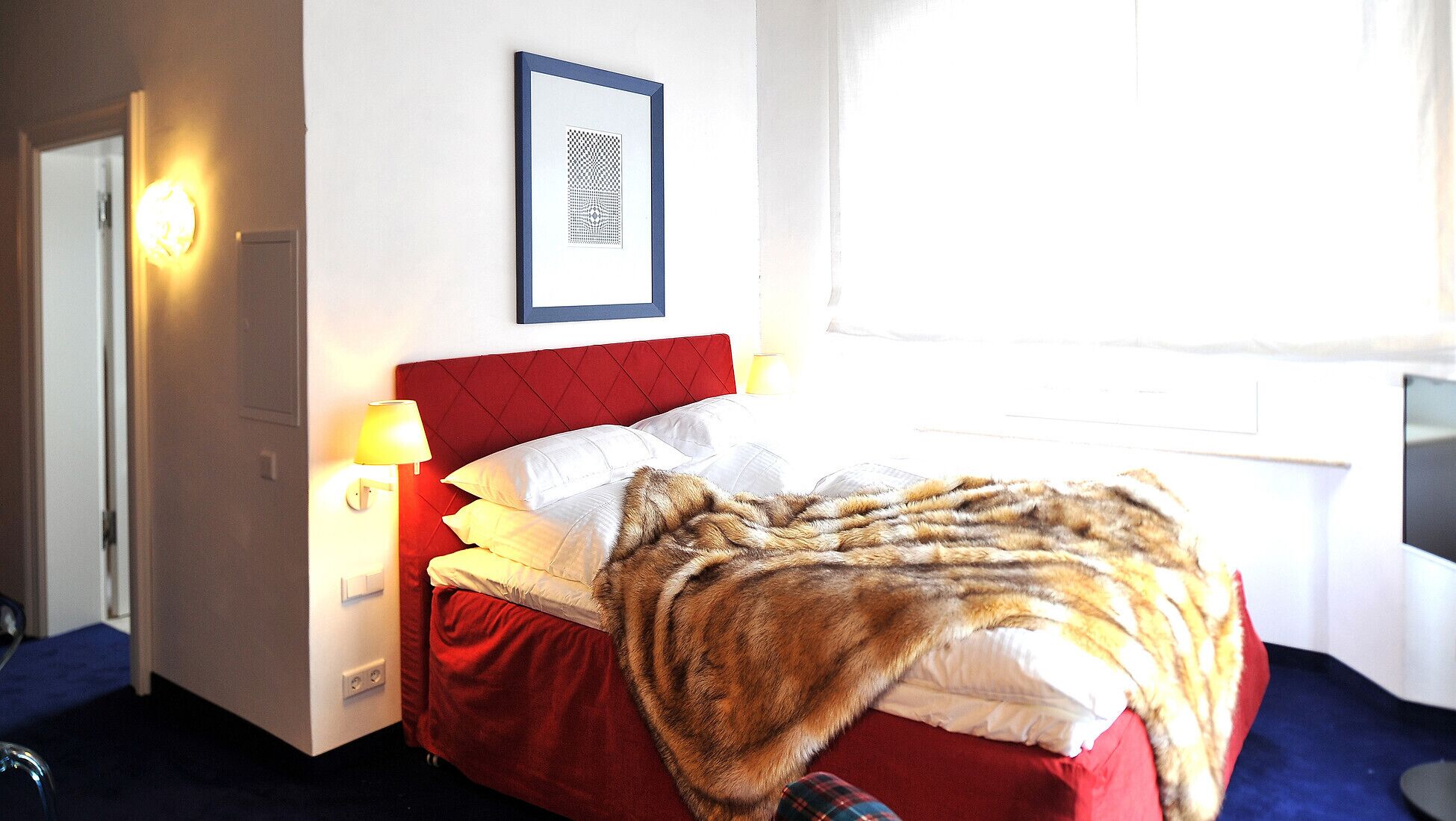 Une chambre d’hôtel avec une tête de lit rouge, du linge de lit blanc, un tableau encadré au mur et une fenêtre laissant entrer la lumière naturelle.