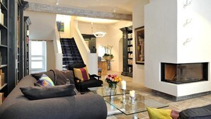 Una sala de estar moderna con una mesa de centro de vidrio, un sofá con cojines de colores, una chimenea y una escalera que sube a un nivel superior.