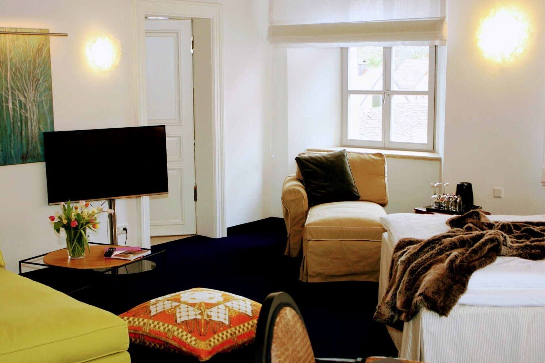 A hotel room with a bed, a sofa, a television, a window, and a small table with a vase of flowers.