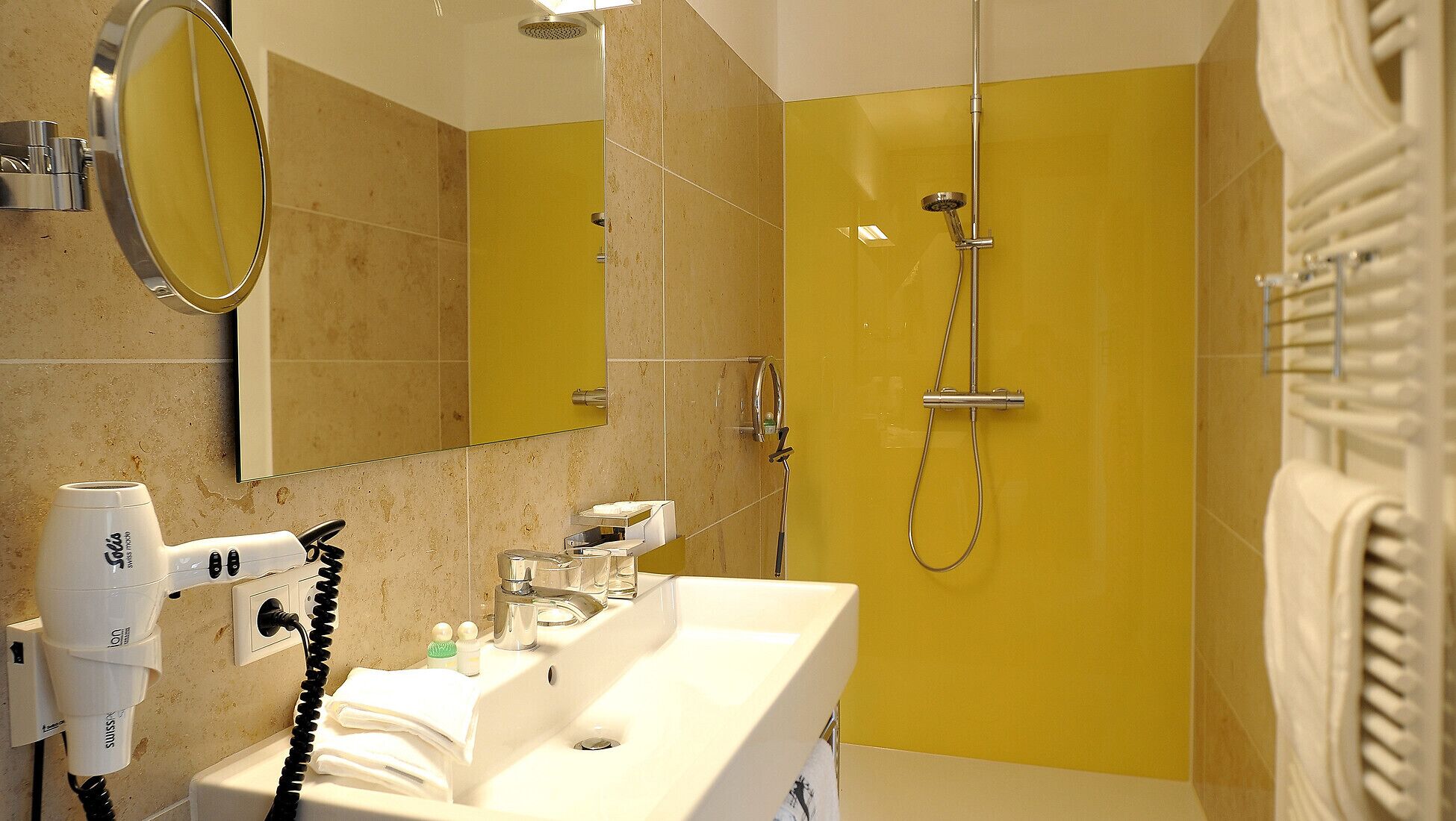 Un baño con una pared amarilla, un lavabo, un espejo, un cabezal de ducha de mano y un perchero para toallas.
