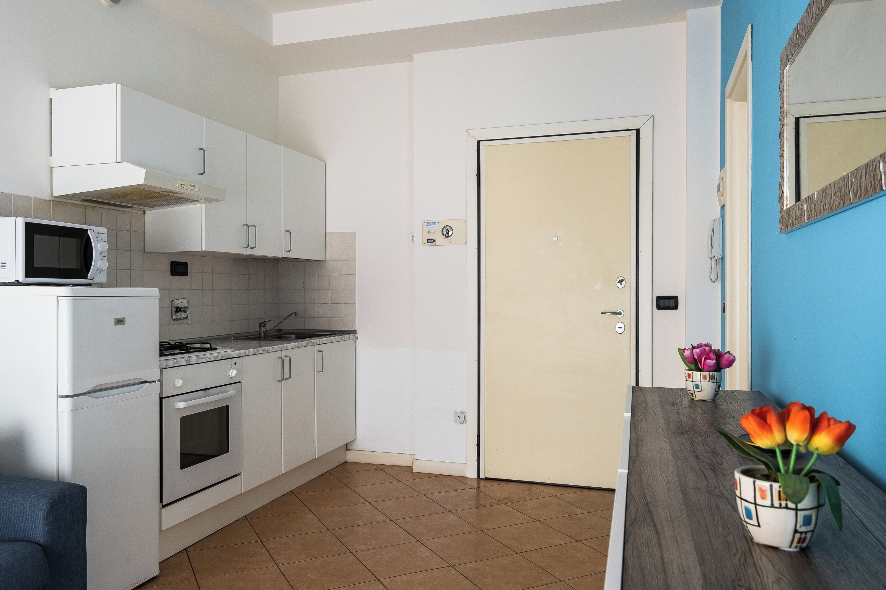 Una cocina con armarios blancos, un microondas, una estufa y un lavabo. Hay una puerta con un interruptor de luz y un teléfono en la pared.