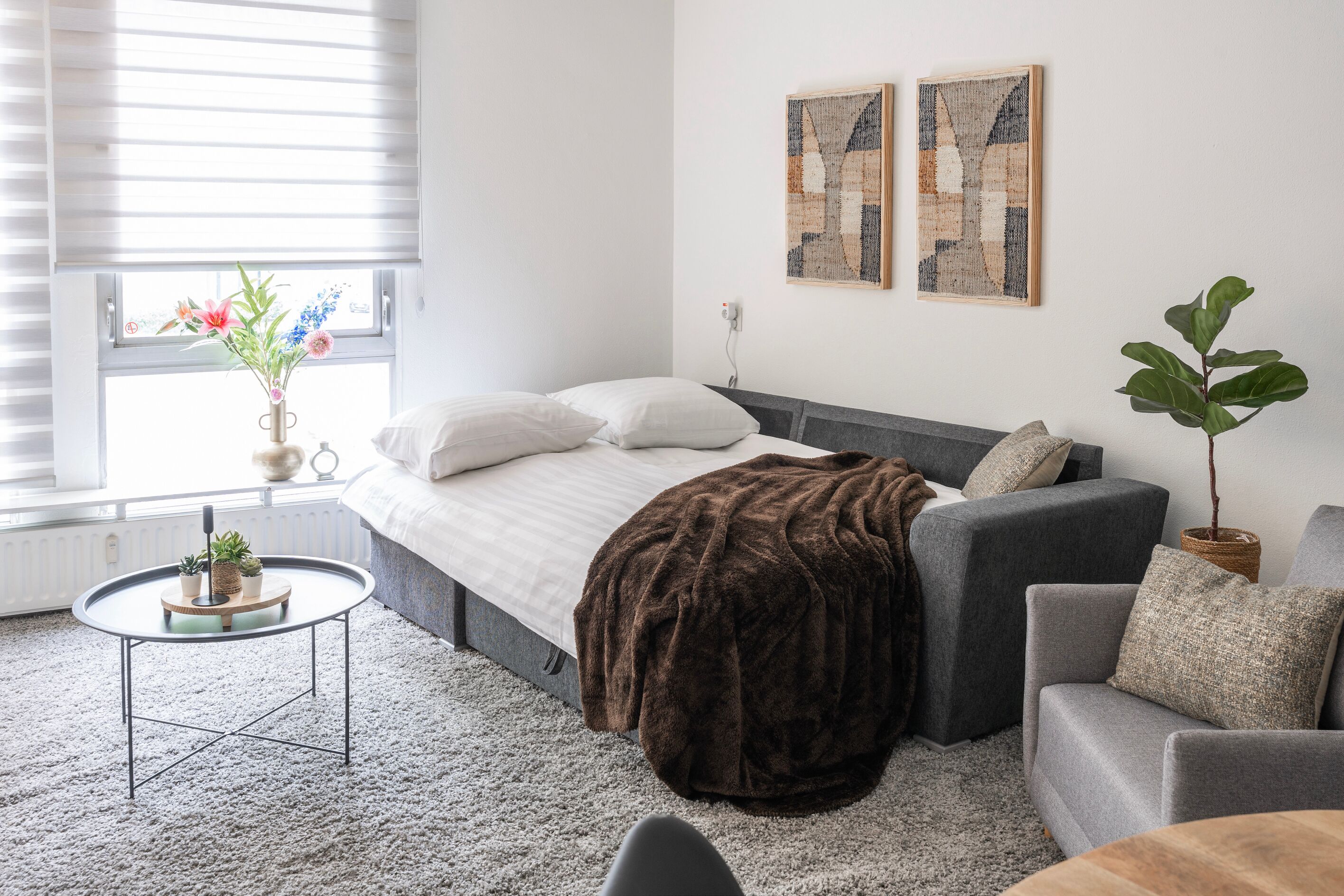 Quarto moderno com cama, sofá cinzento e uma pequena mesa redonda com uma planta.