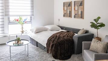 Quarto moderno com cama, sofá cinzento e uma pequena mesa redonda com uma planta.