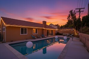 A swimming pool with lounge chairs and string lights, surrounded by a house with a tiled roof and a brick chimney.
