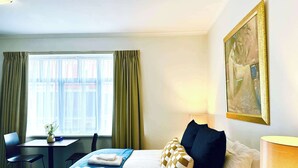 Ein Hotelzimmer mit Bett, Schreibtisch, Stuhl, einem Fenster mit Vorhängen und einem Bild an der Wand.