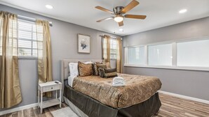 Ein Schlafzimmer mit Bett, Nachttisch, Deckenventilator und einem Fenster mit Vorhängen.