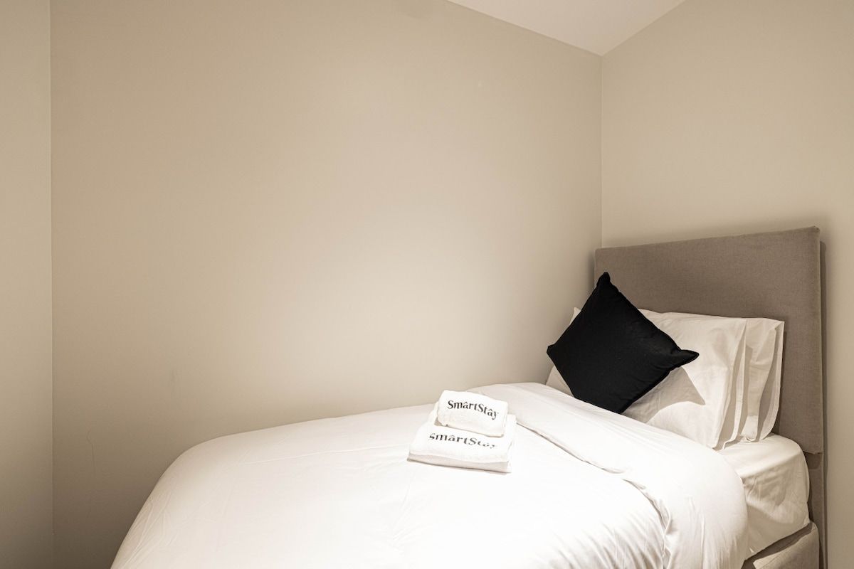 Ein ordentlich bezogenes Bett mit weißen Leinen und einem schwarzen Kopfkissen, ein gefaltetes weißes Handtuch mit 'SmartStay'-Branding und eine schlichte Wand.
