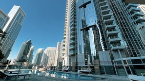 A modern high-rise building with a glass facade, a rooftop pool area with lounge chairs, and a clear blue sky.