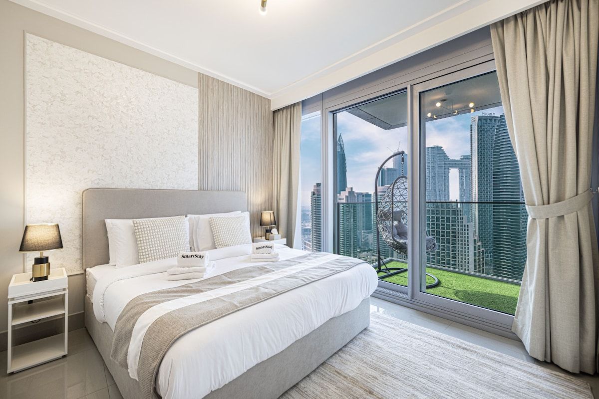 A modern hotel room with a large bed, a nightstand, a lamp, and a view of the cityscape through floor-to-ceiling windows.