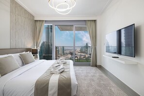 A hotel room with a large bed, a flat-screen TV, a balcony with a city view, and a modern light fixture.
