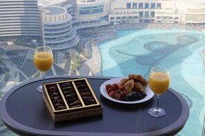 A table with a box of chocolates, a plate of fruits, and two glasses of orange juice, overlooking a cityscape and water.