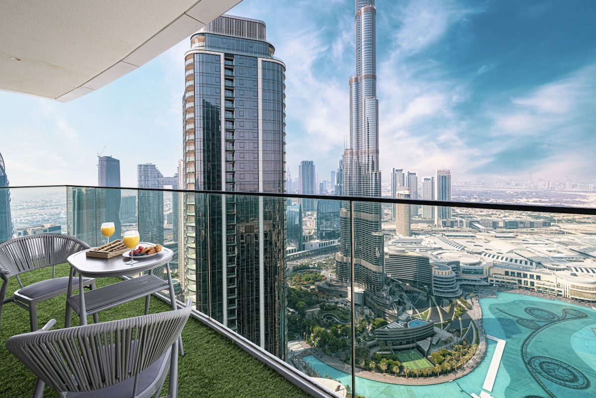 Un balcón con barandilla de vidrio que ofrece vistas a un paisaje urbano, con edificios modernos de gran altura y un área ajardinada con piscina.