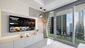 A modern living room with a large flat-screen TV displaying a movie poster, a balcony with a city view, and a well-lit space with a ceiling light. - New 4BR Penthouse | Burj Khalifa View |Opera Grand (Dubai)