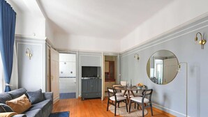 A living room with a grey sofa, a dining table with chairs, a TV, and a kitchen area in the background. - Top Location 4P Studio - Champs-Elysées Best Stay (Paris)