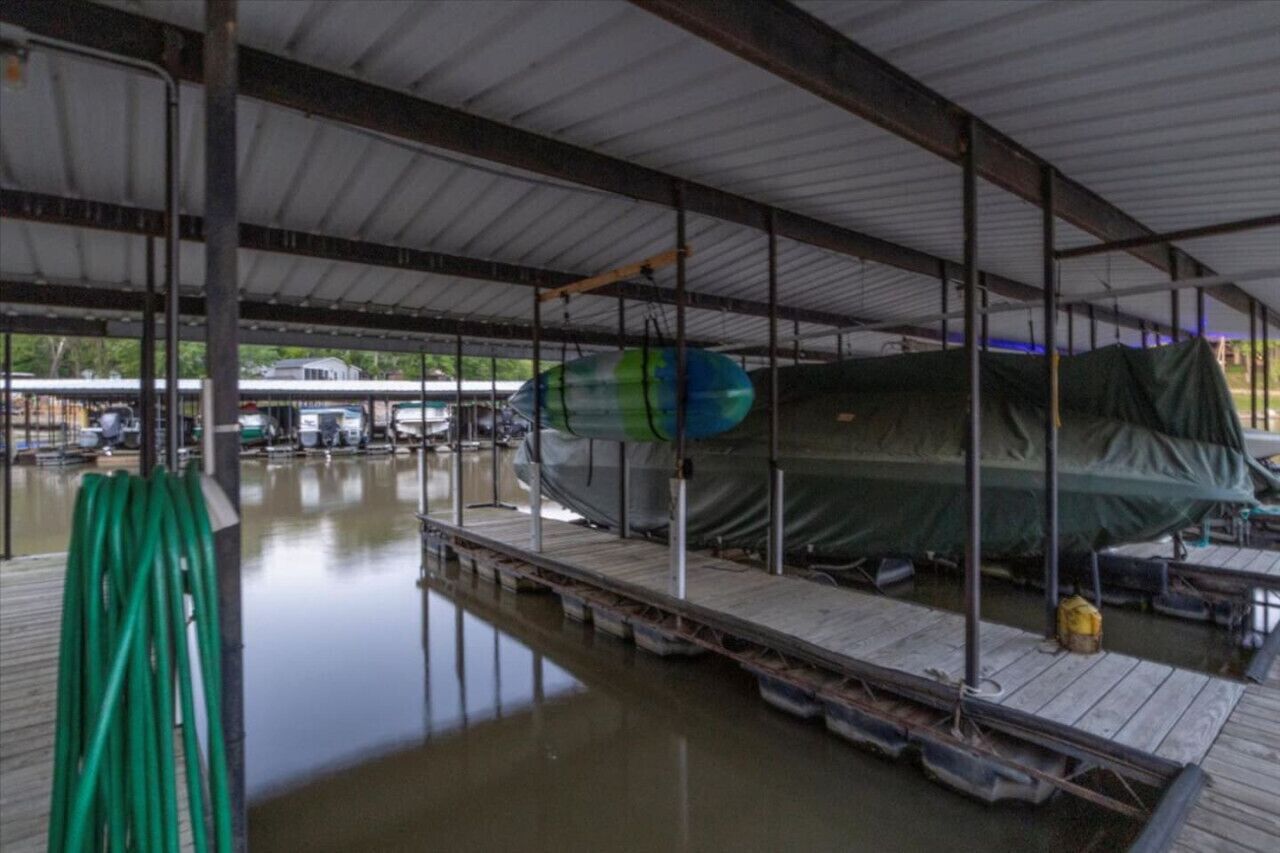 Een overdekte bootopslag met een boot onder een groene zeil, een groene tuinslang en een steiger die in het water uitloopt.