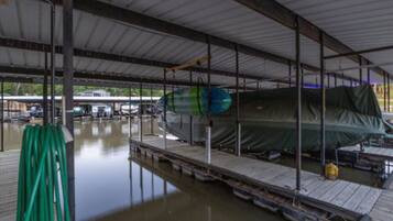 Een overdekte bootopslag met een boot onder een groene zeil, een groene tuinslang en een steiger die in het water uitloopt.