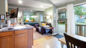 A modern kitchen with a built-in oven, a dining area with a wooden table and chairs, a cozy living room with a sofa and a TV, and a balcony with a view of the backyard.