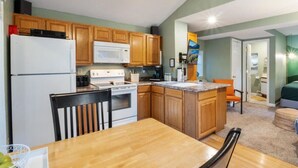 A compact kitchen with wooden cabinets, a white refrigerator, a stove, and a microwave. There is a dining table with chairs and a small island with a sink.