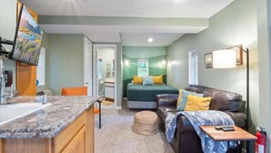 A compact room with a bed, a couch, a small table, a television, and a bathroom visible through an open door.