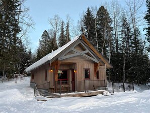 A wooden cabin with a porch and a small deck, surrounded by snow and trees.