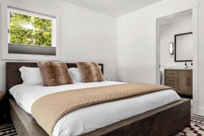 A bedroom with a large bed, a window with a view of trees, a wooden headboard, and a wooden nightstand.