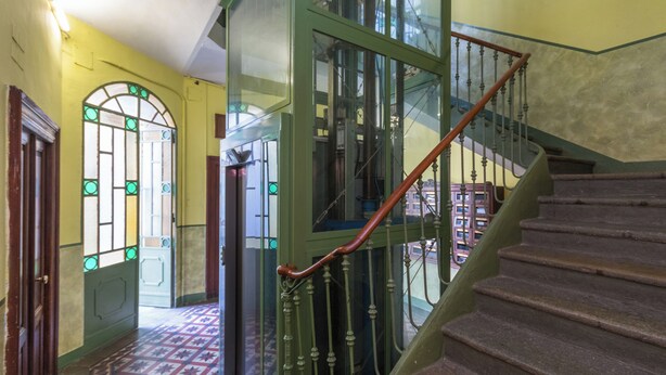 Un escalier avec une rampe en bois et des balustrades en métal vert, menant à une pièce dotée d’un sol à motifs et d’une porte ornée de vitraux.