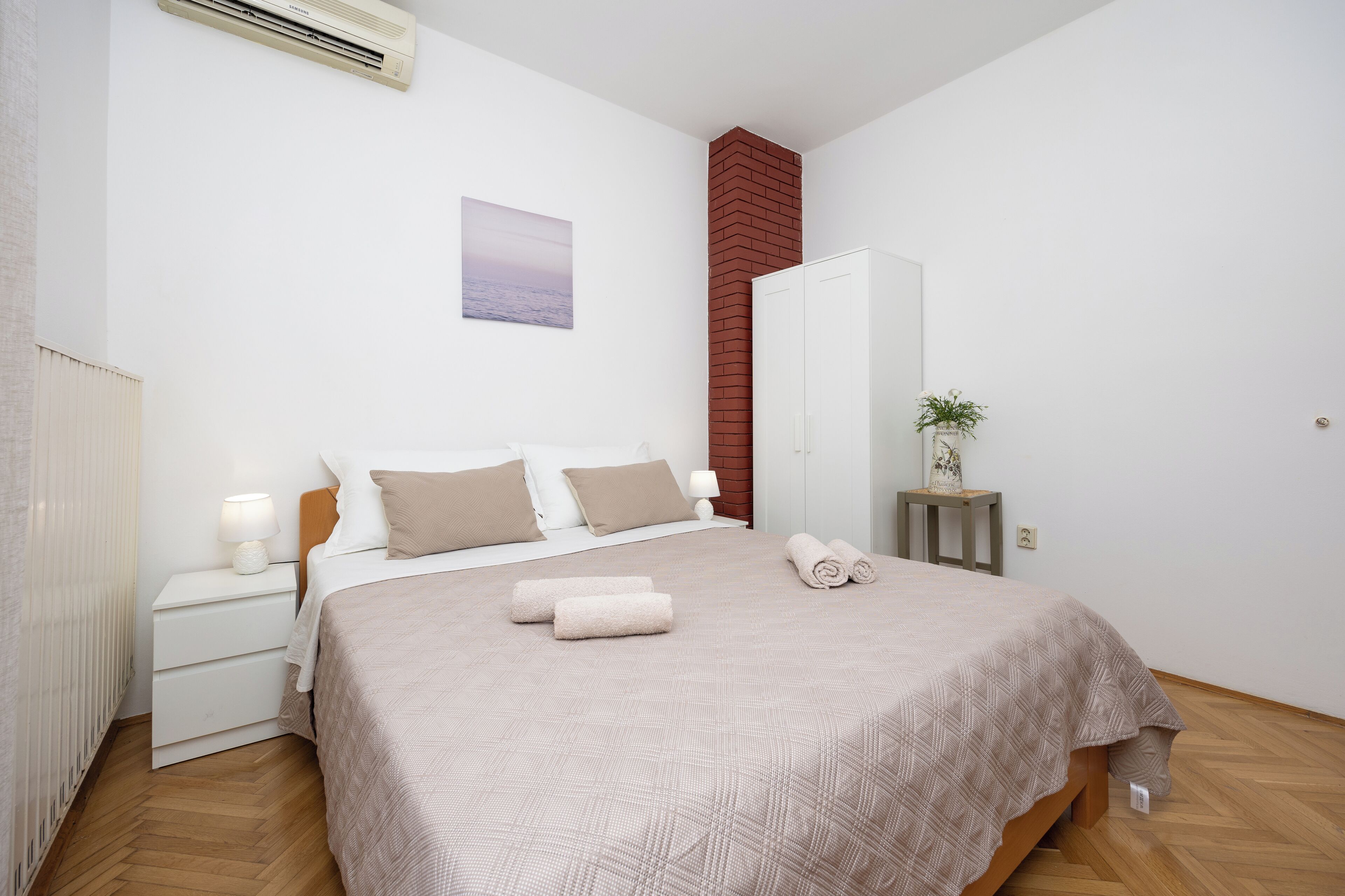 A neatly arranged bedroom with a bed, bedside tables, a wardrobe, and a small plant on a nightstand.