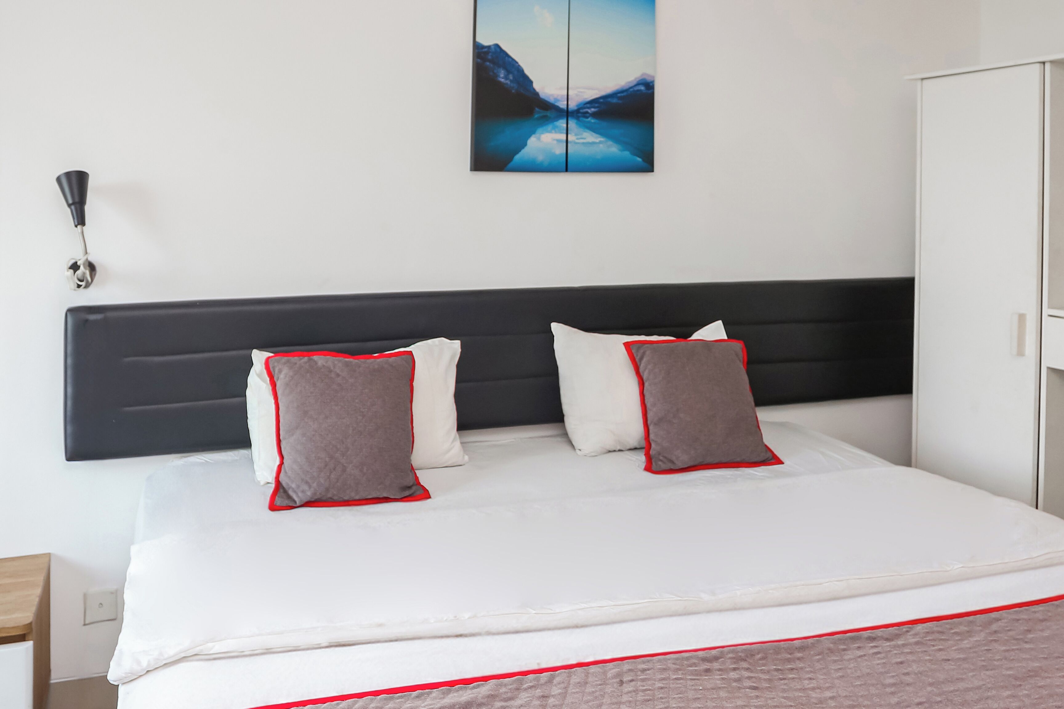 Un lit bien fait, avec une literie blanche et rouge, une tête de lit noire et un tableau représentant un paysage de montagne accroché au mur.