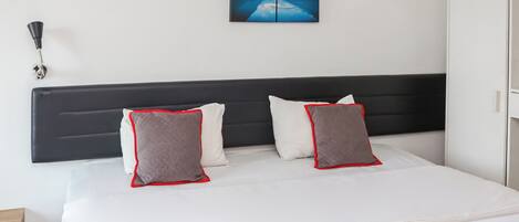 A neatly made bed with white and red bedding, a black headboard, and a painting of a mountain landscape on the wall.