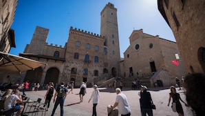 Un edificio histórico con una torre central y ventanas arqueadas, rodeado por un patio con gente y mesas al aire libre.