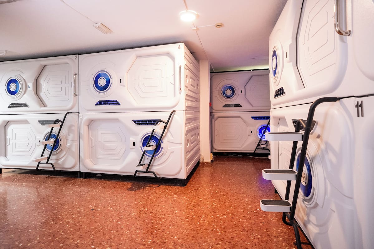 Une rangée de chambres d’hôtel blanches, au design moderne, compactes et bien éclairées, toutes conçues selon un modèle uniforme, avec un panneau de commande central et une petite échelle pour y accéder.