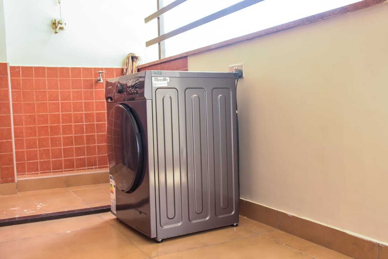 Eine kompakte, moderne Waschmaschine steht in einer Ecke des Raumes, dessen Wände mit Fliesen verkleidet und ein Fenster vorhanden ist.
