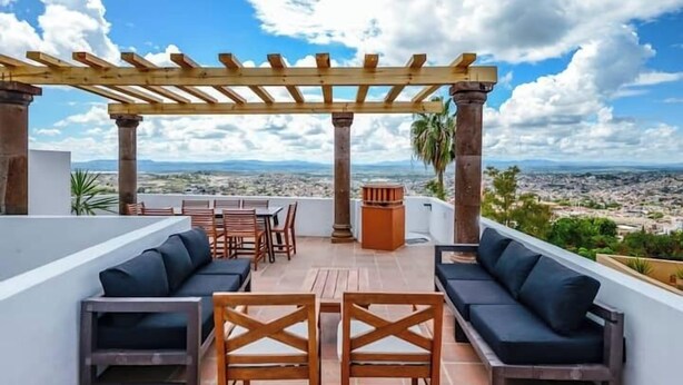 Un patio al aire libre con mobiliario de madera, una pérgola y vistas a un paisaje urbano.